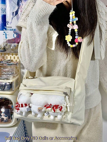 Cream ita crossbody bag worn on shoulder with three white plushie dolls displayed in clear front zip window