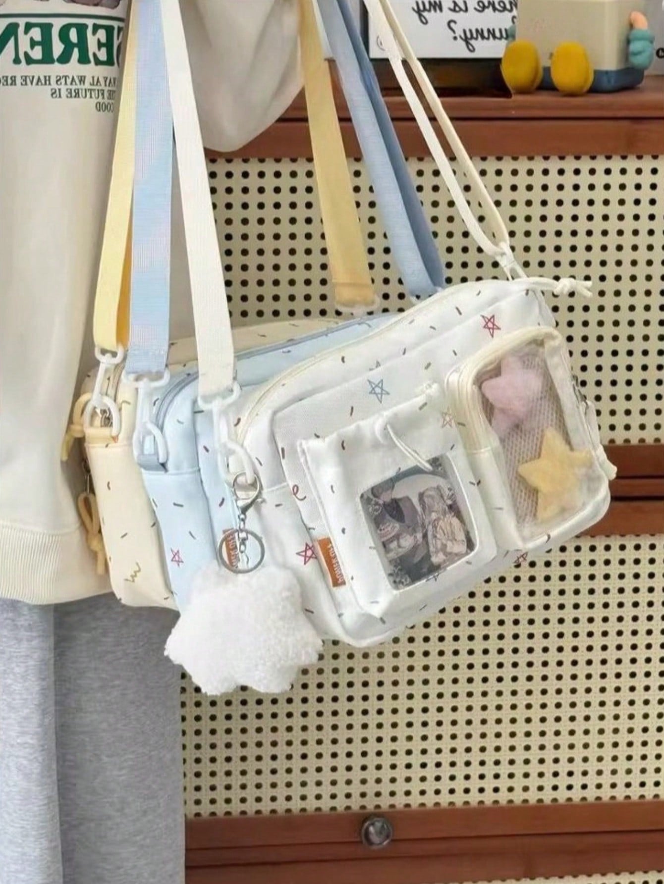 Two confetti crossbody ita bags hanging – cream and pale blue – with plush charms and photocard inserts shown